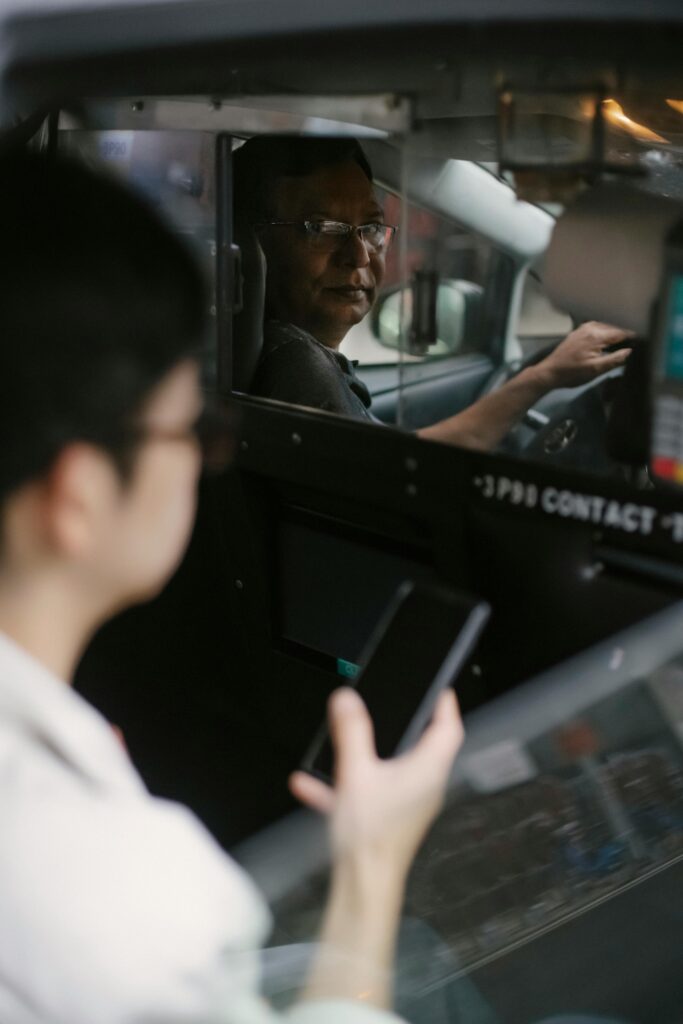 View through open window of ethnic taxi driver looking over shoulder on passenger in glasses surfing smartphone
